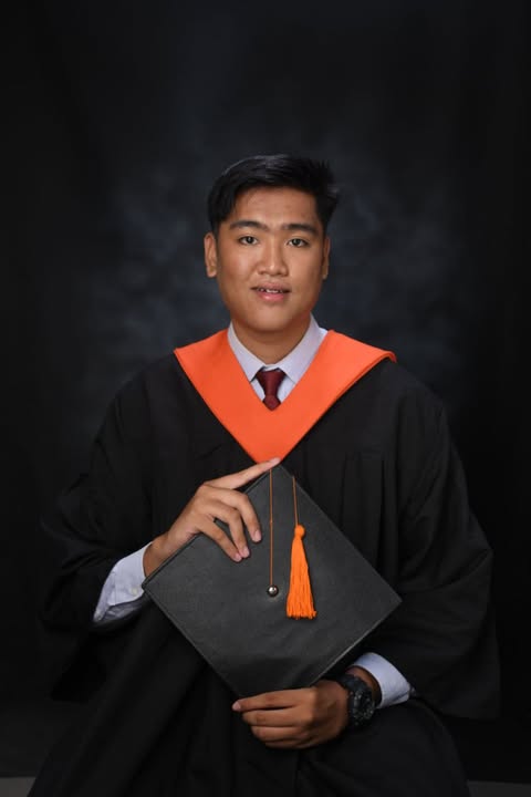 Jorenzon Miguel in a black graduation gown and orange stole, holding a mortarboard with an orange tassel